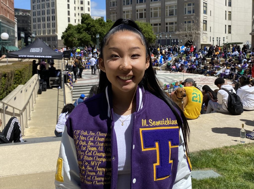 A Thai American girl wearing a purple letterman's jacket with the letter T smiles at camera