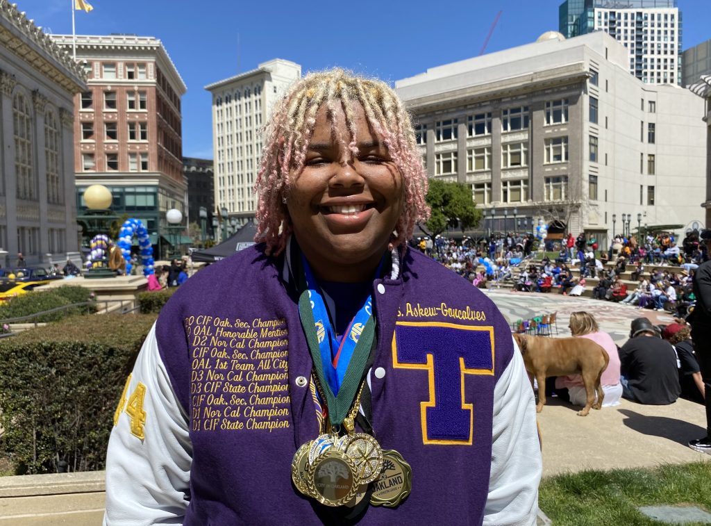 An African American girl with blonde and brown twisted hair wearing a purple letterman's jacket smiles for camera.
