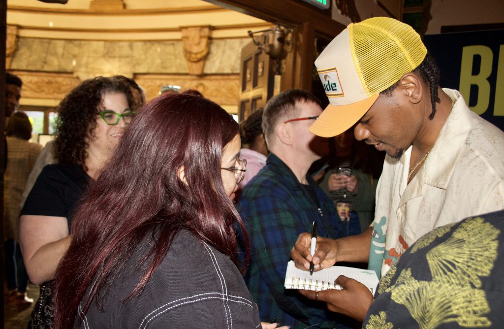 an African American man wearing a yellow trucker hat signs an autograph for a young woman
