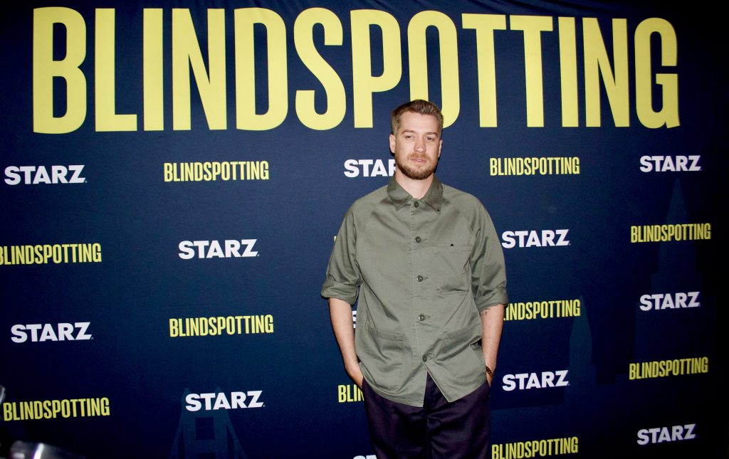 A man with light hair wearing a light green collared shirt poses in front of a black background with "Blindspotting" in yellow letters