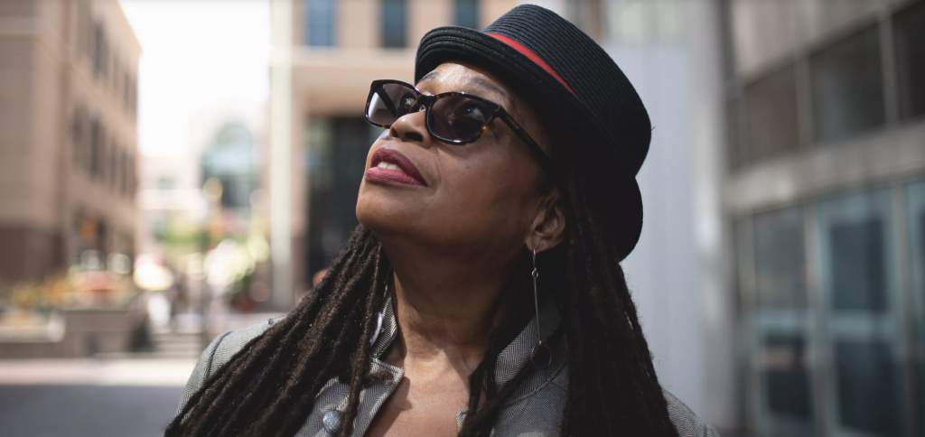 An African American woman looks upwards wearing a hat and sunglasses with a slight smile