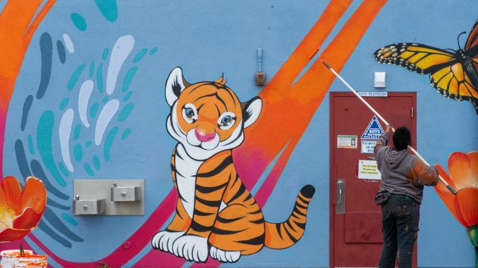 A school wall with a cute baby tiger, and a woman finishing painting an orange line on the mural