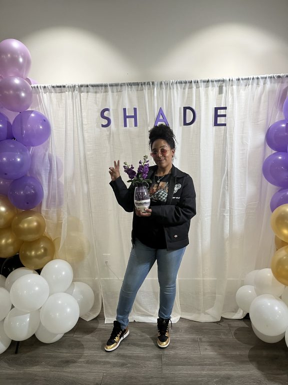 An African American woman stands in front of a photo booth background with the word SHADE and she holds up a peace sign