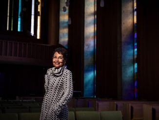 An elder African American woman with short hair and a dress smiles for camera inside a church with colorful windows