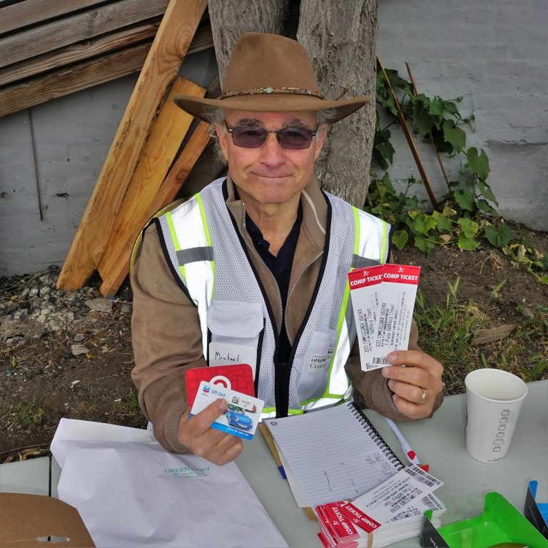 A man wearing sunglasses and a brown hat holds up A's tickets and gift cards whiles letting behind a table outdoors