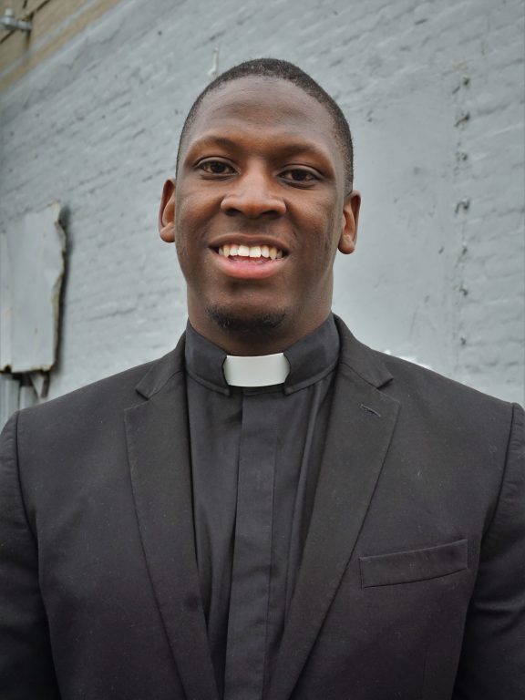 An African American young male minister with short hair wearing a black suit stands for a portrait, smiling