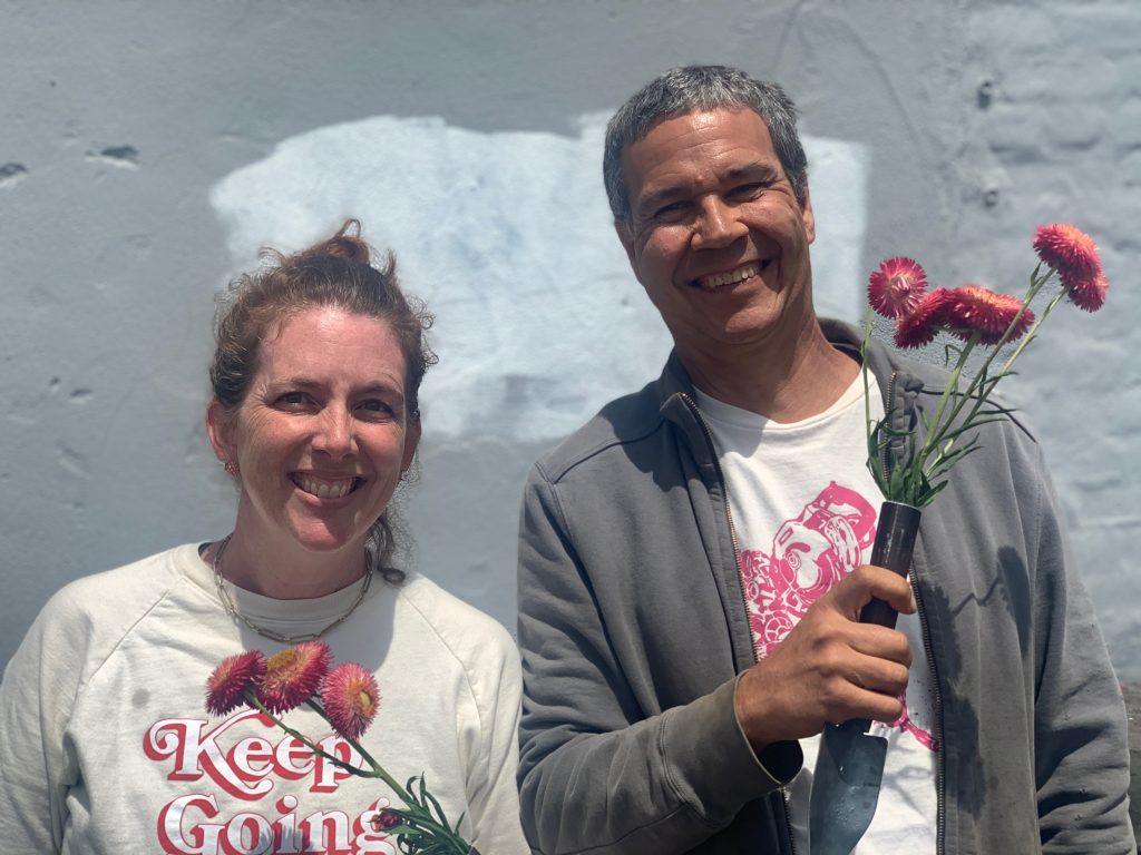 a white woman in a t-shirt stands next to a man with short grey hair holding a vase of flowers
