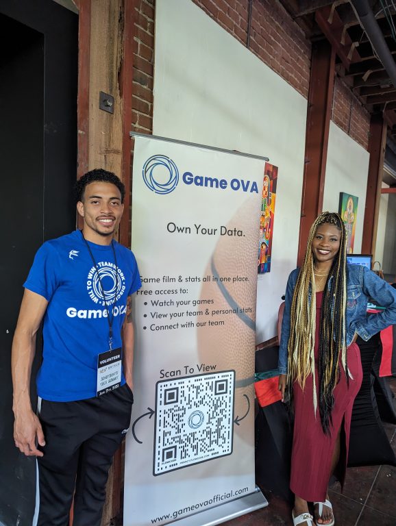 A young African American man and an African American woman stand next to tall banner that says Game Ova