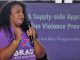 An African American woman with curly hair holding a mic speaks in front of a purple display about gun violence
