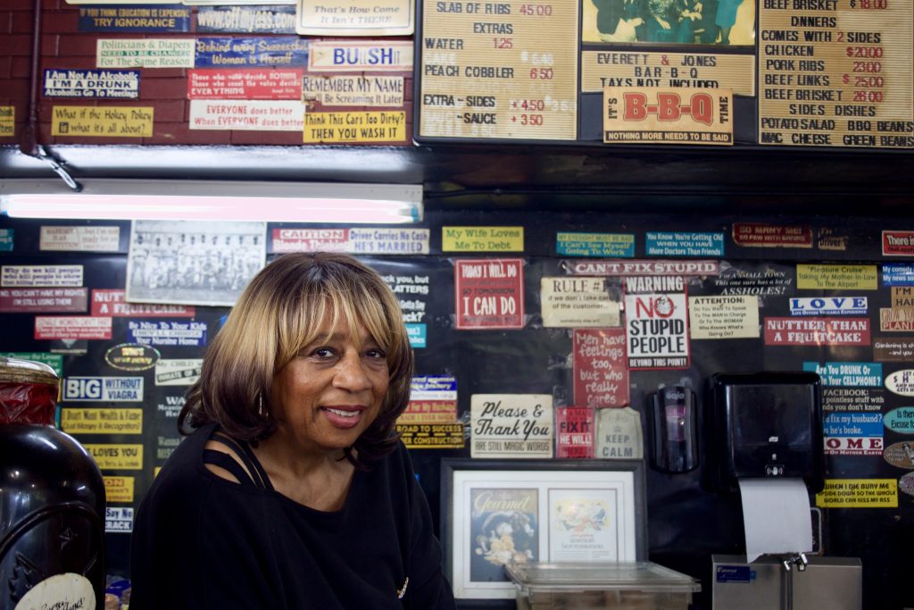 A Black woman leans against a counter in a bbq restaurant, with a lot of tongue in cheek stickers on the wall behind her