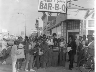 a black and white photo of a storefront that says Everett & jones Bar-B-Q and a group of Black people dressed in 70s attire pose in front of it