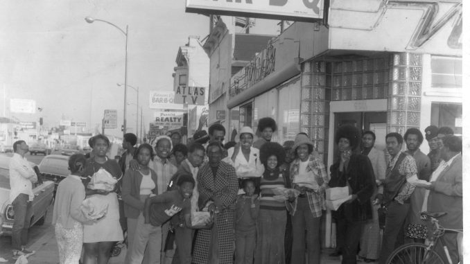 a black and white photo of a storefront that says Everett & jones Bar-B-Q and a group of Black people dressed in 70s attire pose in front of it