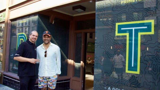 A man with a low pony tail and polo shirt stands next to an African American man in a white shirt in front of a shop that has the letter P and T in large lettering