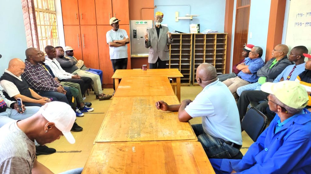 An elder Black man wearing a colorful hat presents to a group of Black men.