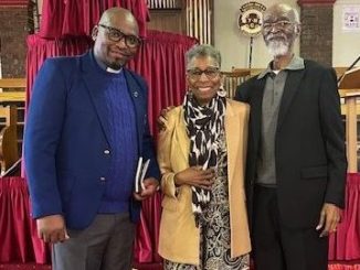 A Black pastor, and elder African American woman, and elder African American man pose for a photo inside a church with red carpet