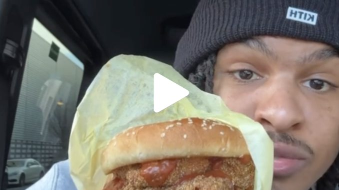 A man sits inside a car and shows the camera a fried fish sandwich