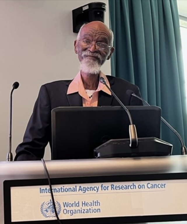 An elder African American man speaks behind a podium that has a World Health Organization branding on it