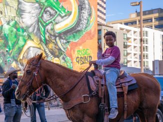 A young Black boy sits on a horse and shows a peace sign with his hand