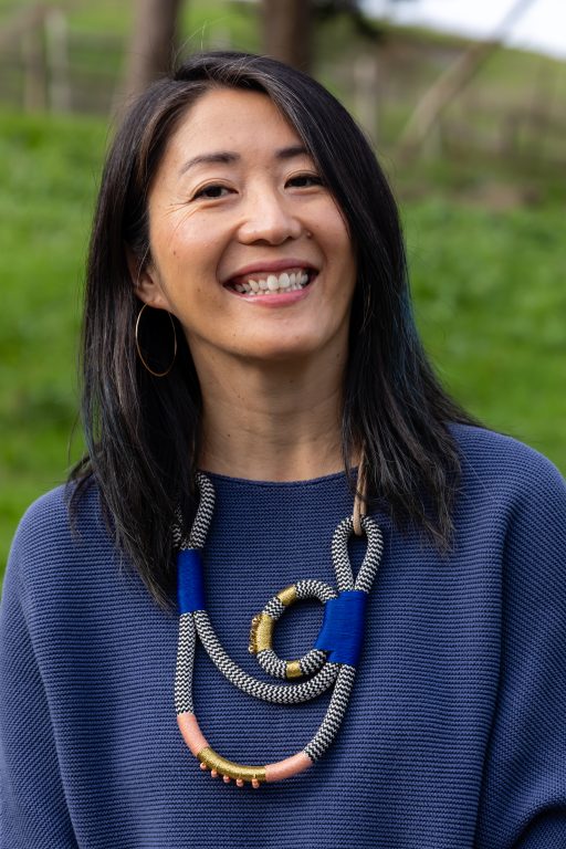 A Chinese American woman wearing a big necklace and royal blue shirt smiles outdoors