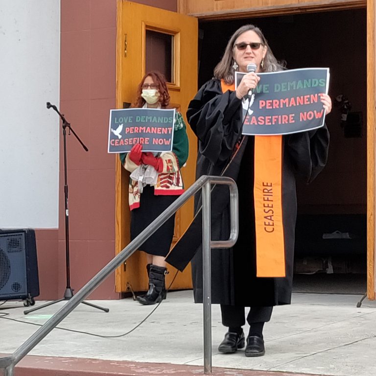 A white female reverend stands in front of a church with a microphone holding a ceasefire sign