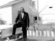 a black and white photo of a Black man in a suit in front of a house